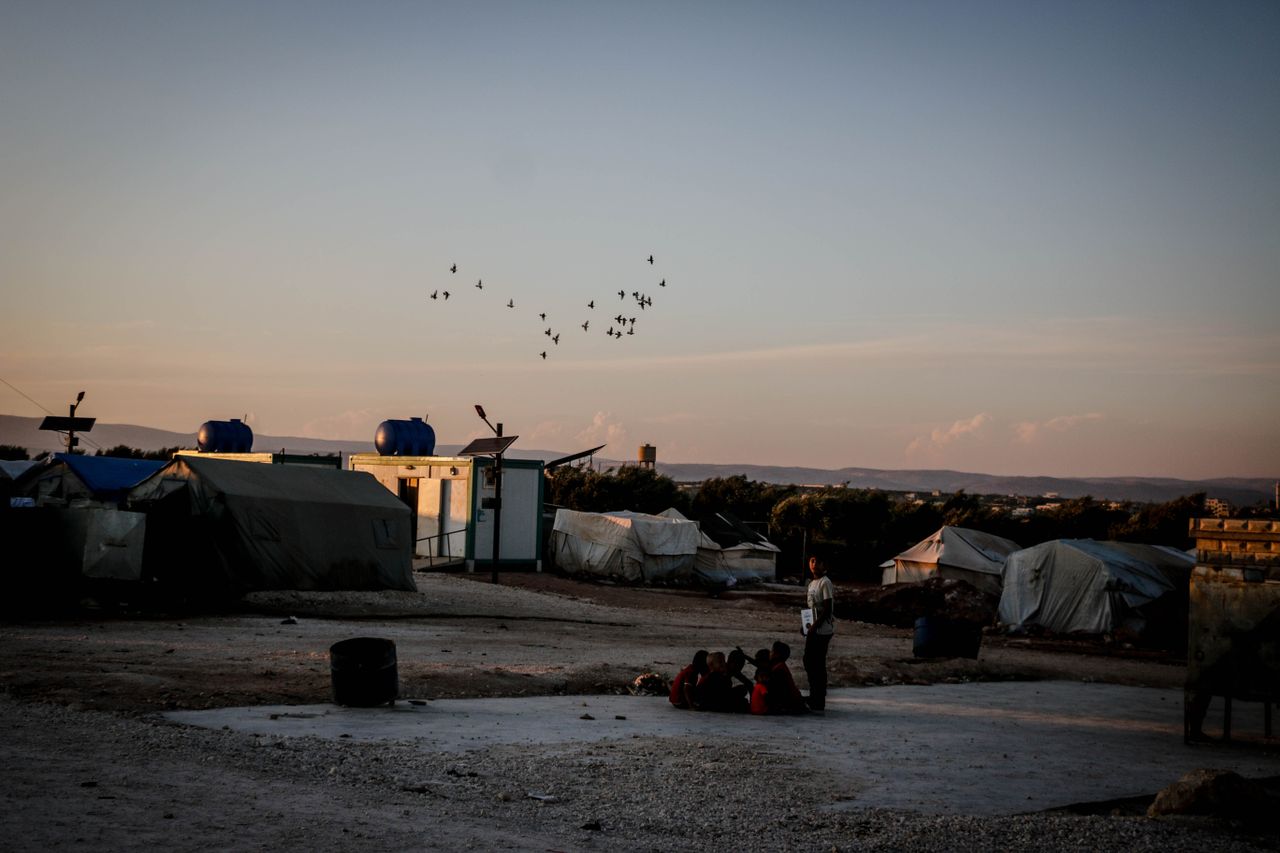 hero-img-02 A group of people in a refugee camp in Syria, with birds flying overhead at sunset.