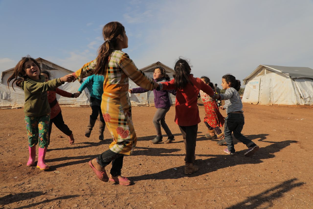 hero-img-01 Children joyfully play outdoors in a Syrian refugee camp, forming a lively circle.