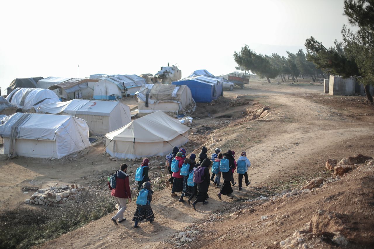 Children carrying backpacks walk through a rural refugee camp under a clear sky.